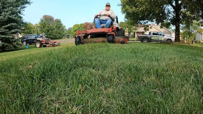 Crew member on a red zero-turn mower in a long-grass yard